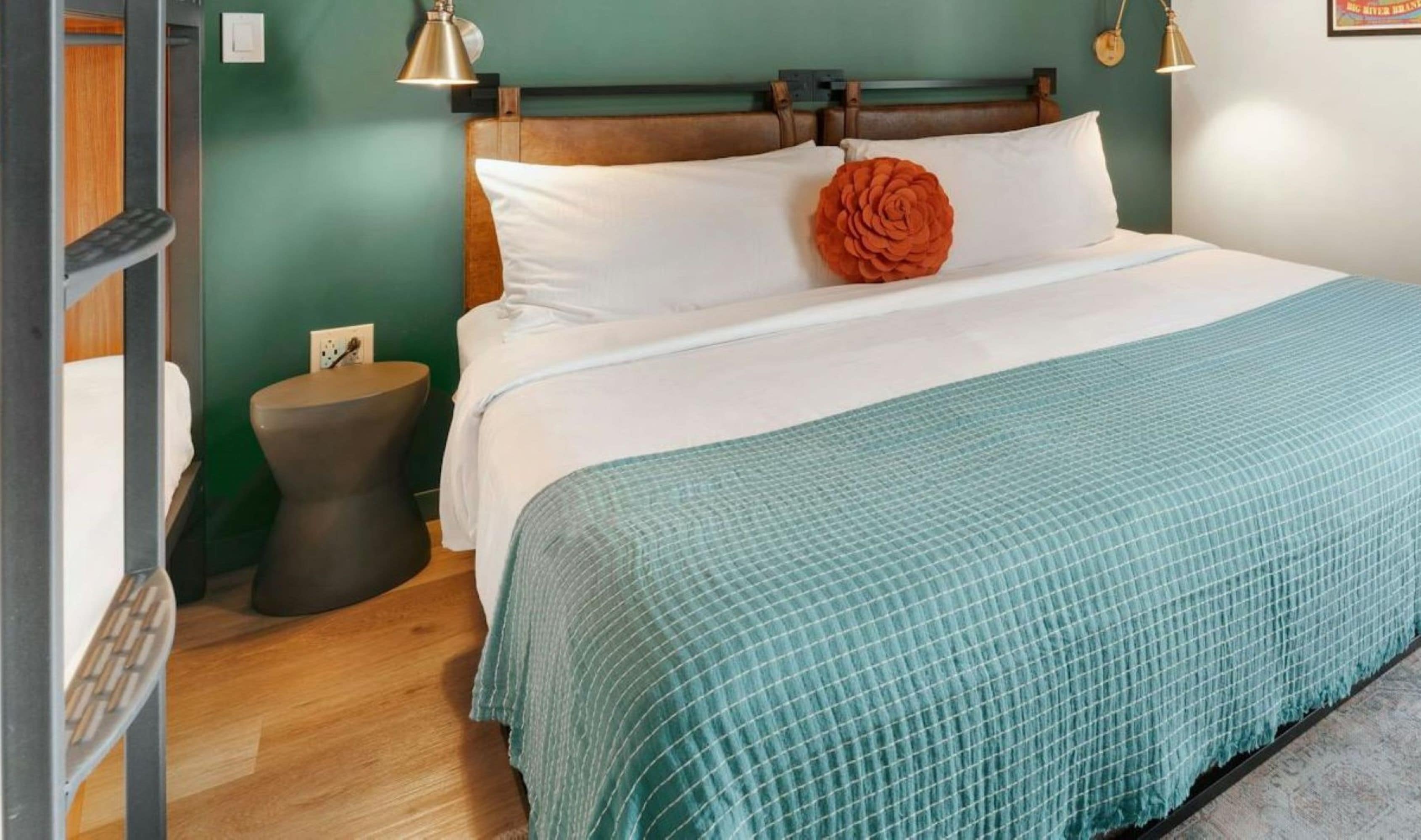 A neatly made bed with a teal checkered bedspread and a large orange flower pillow, a bedside table with a lamp, and a green wall.