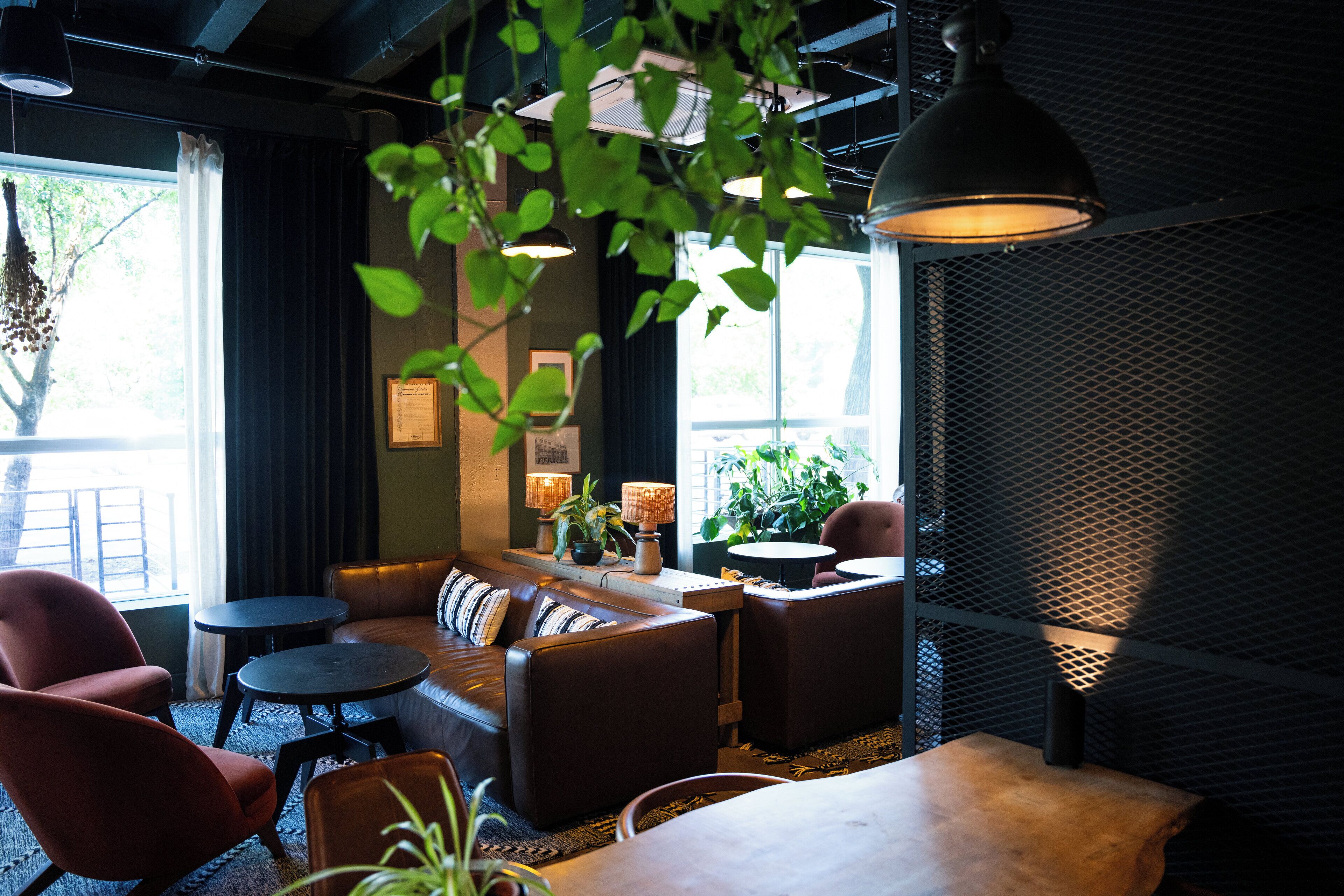A cozy lounge area with a sofa, armchairs, and a table, featuring a large window with natural light and a hanging plant.
