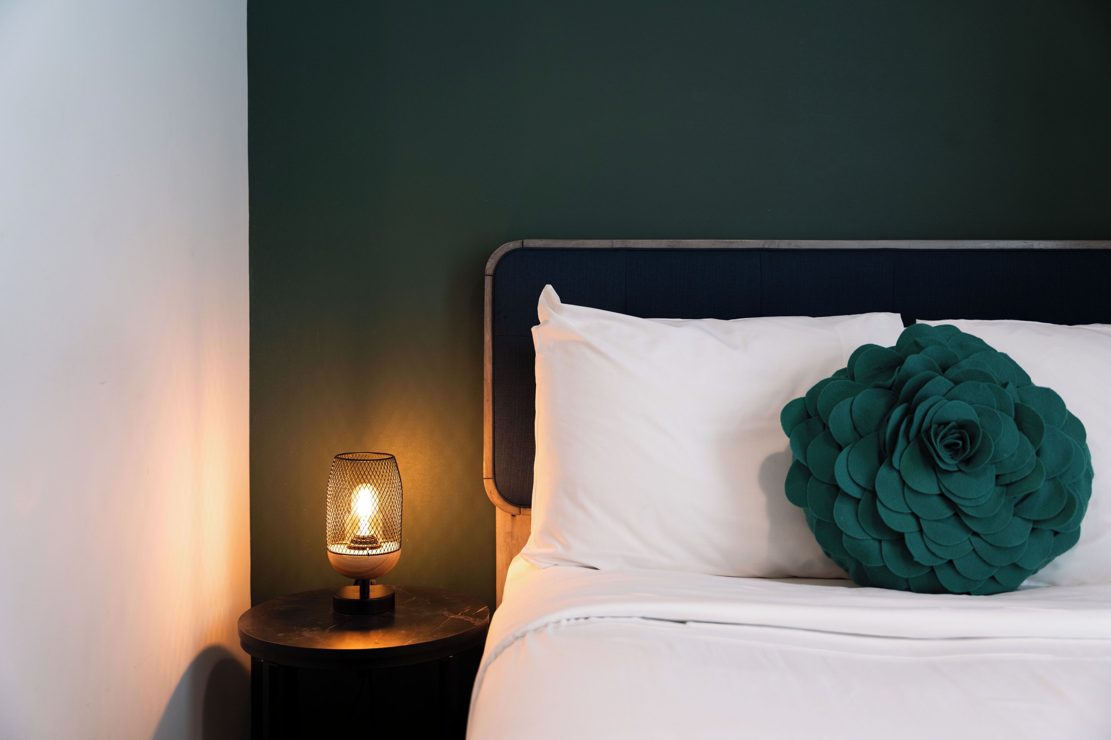 Cama bem arrumada com lençóis brancos e um grande travesseiro decorativo verde, mesa de cabeceira redonda com abajur aceso e cabeceira azul escura contra uma parede verde.