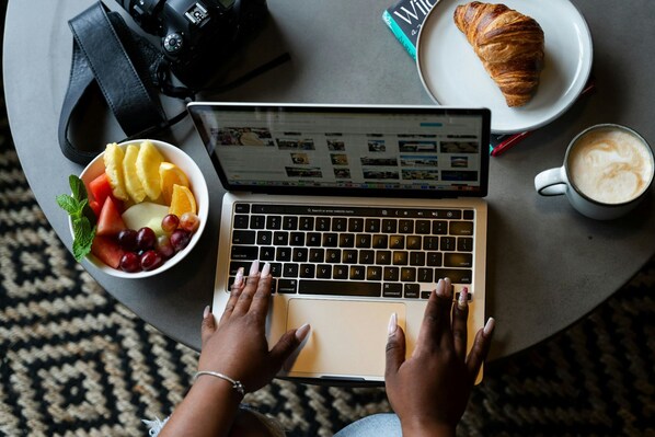 A laptop on a round table with a bowl of fruit, a croissant, and a cup of coffee.