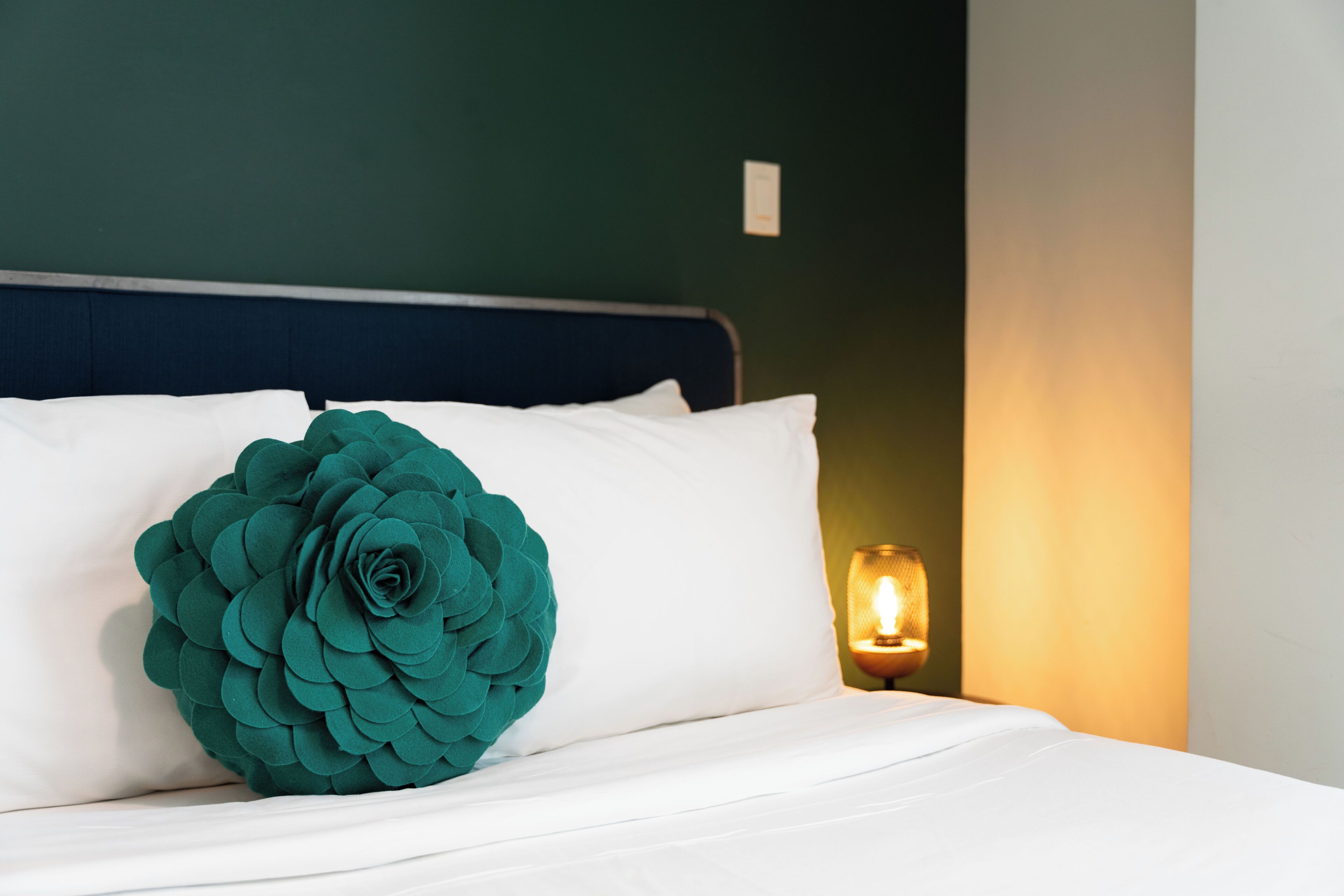 Una cama bien tendida con sábanas blancas y una almohada decorativa color turquesa. Se ve una mesita de noche con una lámpara.