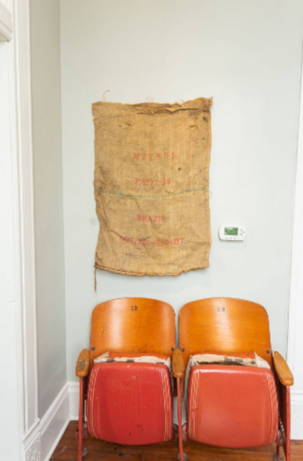 Zwei rote Vintage-Stühle mit Holzlehnen vor einer weißen Wand, ein großes Stück Jutegewebe mit Text und Zahlen, und ein Thermostat an der Wand.