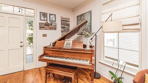 A grand piano in a room with wooden floors, a large window with blinds, and a door with glass panels. - Maison Rose - Midcity (Baton Rouge)