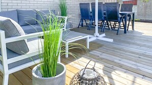 Um terraço com um sofá branco, uma mesa pequena e cadeiras. Há plantas em vasos e um candeeiro no terraço.