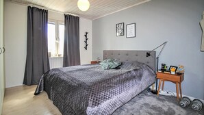 Quarto com cama grande, mesa de cabeceira com abajur, pequeno criado-mudo e um ursinho de pelúcia. Cortinas na janela, relógio de parede e um quadro com uma frase na parede.