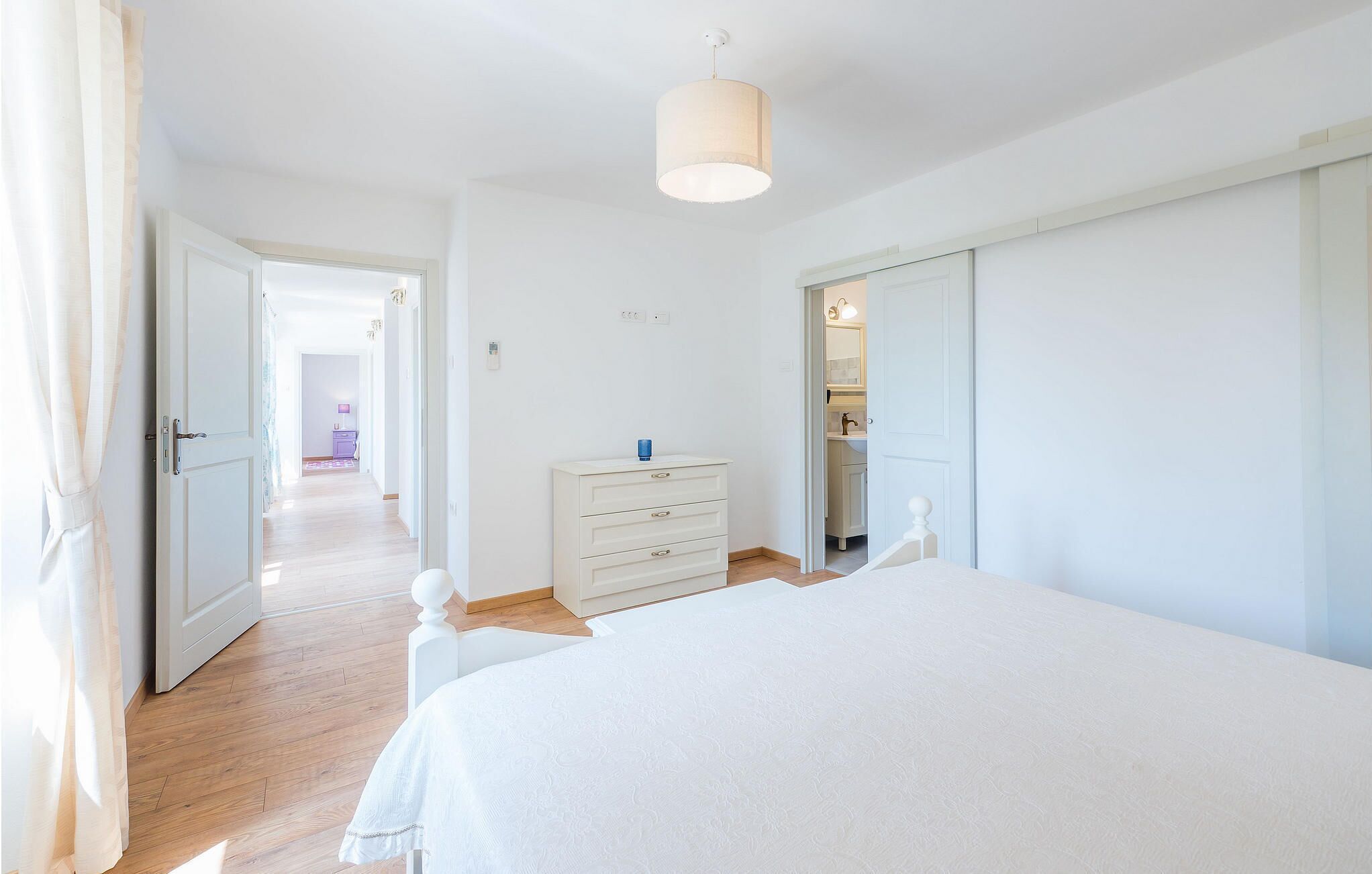 Une chambre avec un grand lit, une commode et une armoire. Un couloir mène à une autre pièce où se trouve une lampe violette.