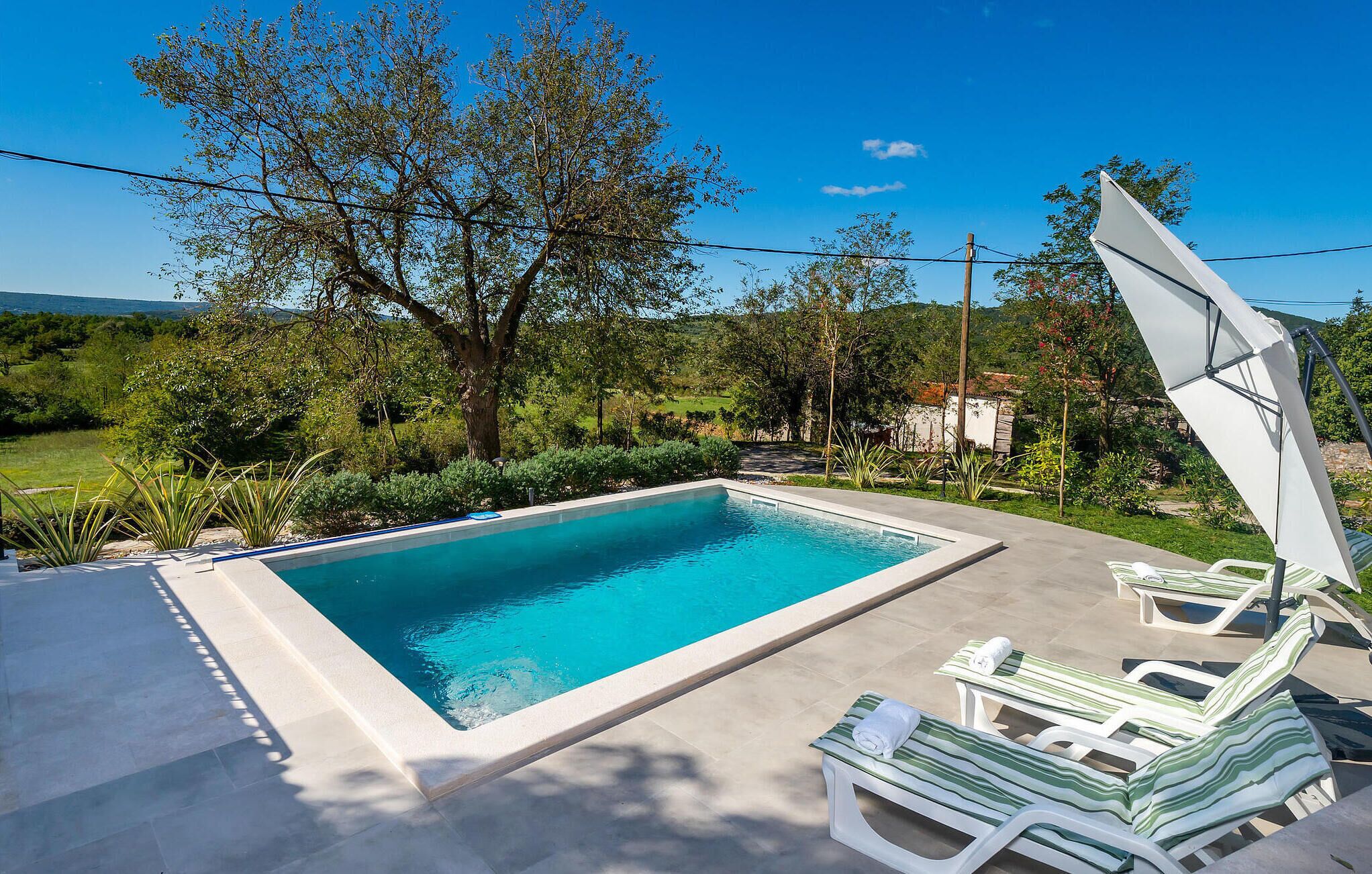 Uma piscina retangular com água azul cristalina, rodeada por um deck de concreto. Há espreguiçadeiras e um guarda-sol branco no deck.