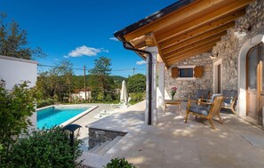 Un espace aménagé au bord de la piscine, avec un mur en pierre, une terrasse en bois et des sièges d’extérieur.