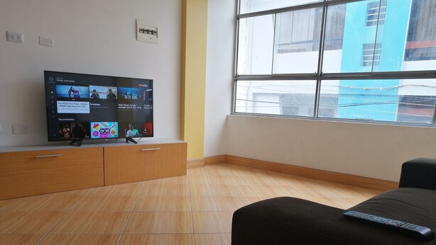 Una sala de estar moderna con un televisor de pantalla plana montado en un mueble de madera, un sofá negro y un ventanal con vista a un edificio azul.