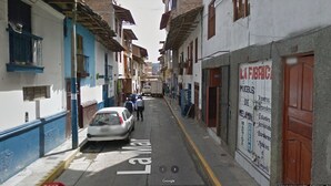 Una calle estrecha flanqueada por edificios, con un coche estacionado y algunos peatones.