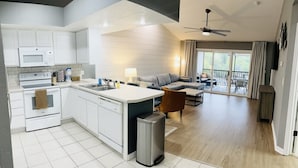 A modern kitchen with white cabinets, a microwave, and a stove. There is a dining area with a gray sofa and a small table. The room has a large window and a ceiling fan.