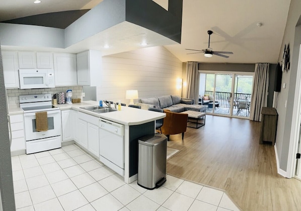A modern kitchen with white cabinets, a microwave, and a stove. There is a dining area with a gray sofa and a small table. The room has a large window and a ceiling fan.