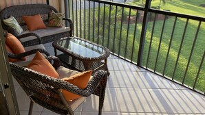 Varanda com móveis de vime, uma mesa de vidro e vista para um jardim bem cuidado e casas.