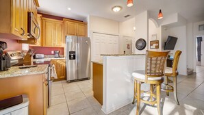 Une cuisine moderne avec des meubles en bois, un réfrigérateur en acier inoxydable, un îlot central intégré avec des tabourets de bar et un éclairage suspendu.