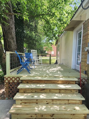 Une terrasse en bois avec des marches menant à un espace abrité, une chaise blanche et une chaise pliante bleue.