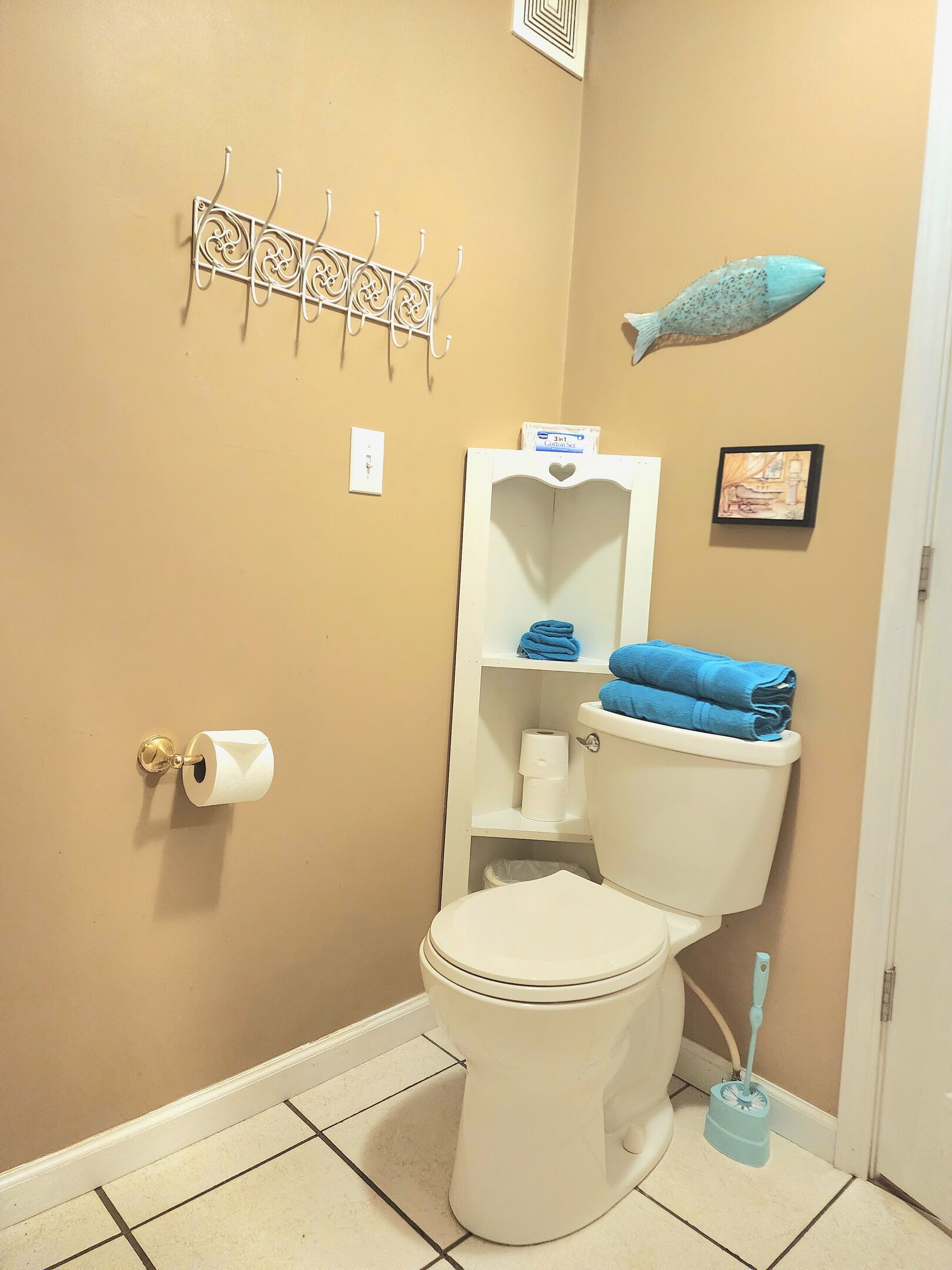 Un baño con inodoro, toallero, dispensador de papel higiénico, adorno de un pez azul y un cuadro en la pared.
