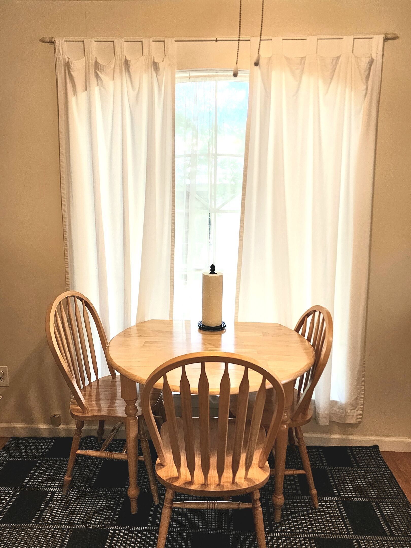A dining area with a round wooden table, four wooden chairs, a candle, and white curtains.