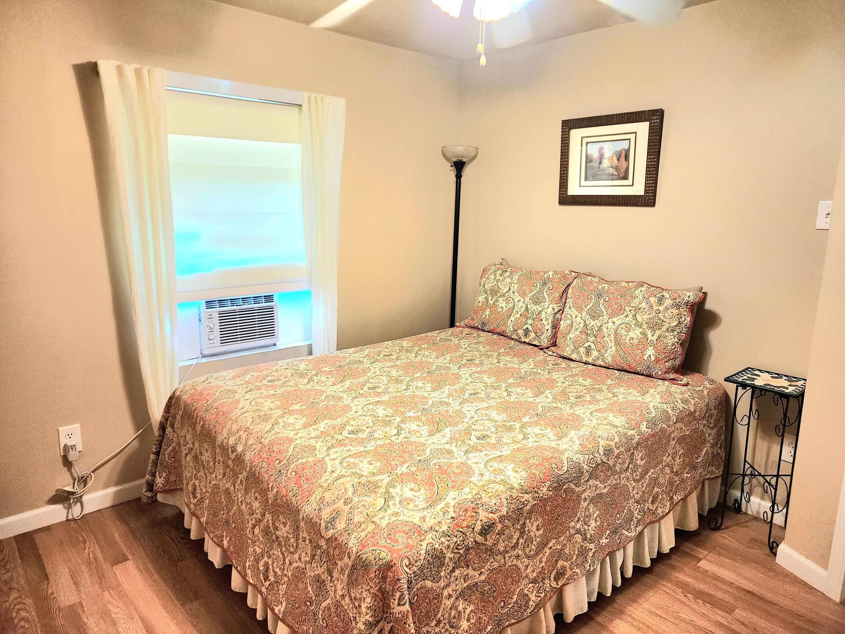 A bedroom with a bed, a window with curtains, a floor lamp, a small table, and a framed picture on the wall.