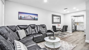 Ein modernes Wohnzimmer mit einer Ledersofa, einem Esstisch mit Stühlen, einem runden Couchtisch, einem großen Spiegel und einer im Hintergrund sichtbaren Küche.