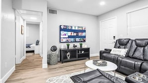 Ein modernes Wohnzimmer mit einer schwarzen Ledersofa, einem weißen Couchtisch und einem an der Wand montierten Fernseher, der verschiedene Sender anzeigt.