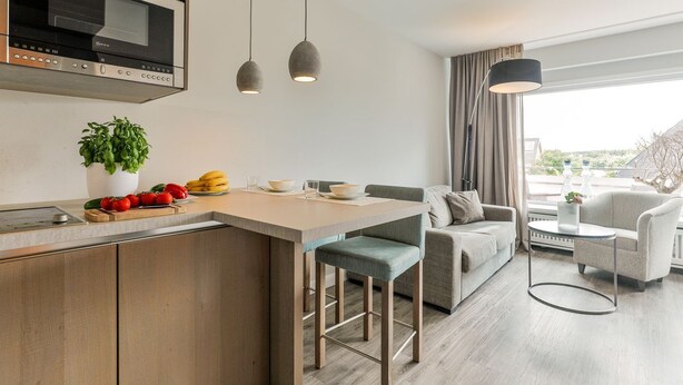 Eine moderne Küche mit einer zentralen Kochinsel, ein Essbereich mit einer Couch und Stühlen sowie ein Blick ins Freie durch große Fenster.