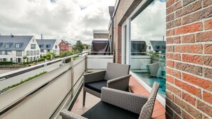 Ein Balkon mit Korbmöbeln, einer Glastrennwand und Blick auf Nachbarhäuser und ein Schwimmbad.