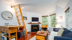 A living room with a fireplace, a TV, a wooden ladder, a couch, and a dining area with a table and chairs. - ❄︎The Nest❄︎ | 1 BR 5 Min Walk to Creekside Gondola | Mountain Rustic Chic (Whistler)