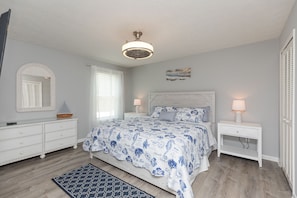 Um quarto com cama, mesas de cabeceira, cômoda, ventoinha no teto, janela com persianas e um tapete.