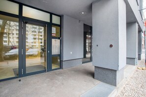 Une entrée d’immeuble avec une porte en verre, un ascenseur en métal et un mur gris à texture.