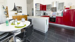 Una cocina moderna con una isla central, armarios rojos y blancos, un horno empotrado y un comedor con mesa blanca y sillas negras.