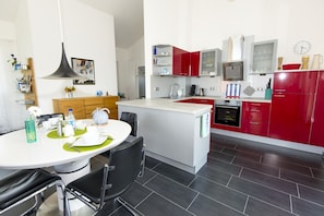 Una cocina moderna con una isla central, armarios rojos y blancos, un horno empotrado y un comedor con mesa blanca y sillas negras.
