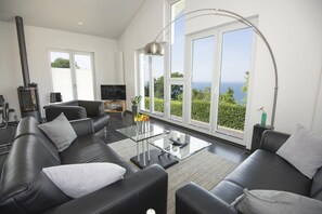 A modern living room with a black leather sofa, a glass coffee table, and a large window offering a view of the sea. - Beautiful sea view from the roof terrace - Haus Ostseeblick 1 FeWo Mee(h)rblick - (Lohme)