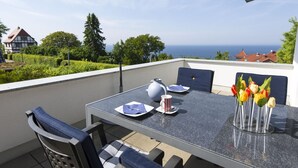 Un balcón con una mesa dispuesta para dos, sillas, un jarrón de tulipanes y vista al mar y casas al fondo.