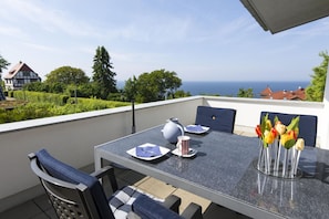 Un balcón con una mesa dispuesta para dos, sillas, un jarrón de tulipanes y vista al mar y casas al fondo.