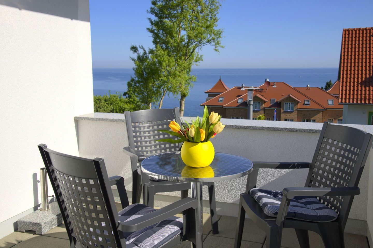 Ein Balkon mit einem runden Tisch und vier Stühlen, einer gelben Vase mit Blumen und Blick auf das Meer sowie Gebäude im Hintergrund.