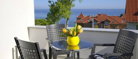 Un balcón con una mesa redonda y cuatro sillas, un jarrón amarillo con flores, y vista al mar y edificios al fondo.