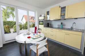 Une cuisine moderne avec des meubles de couleur jaune, une table à manger blanche et une vue sur une maison, visible à travers de grandes fenêtres.