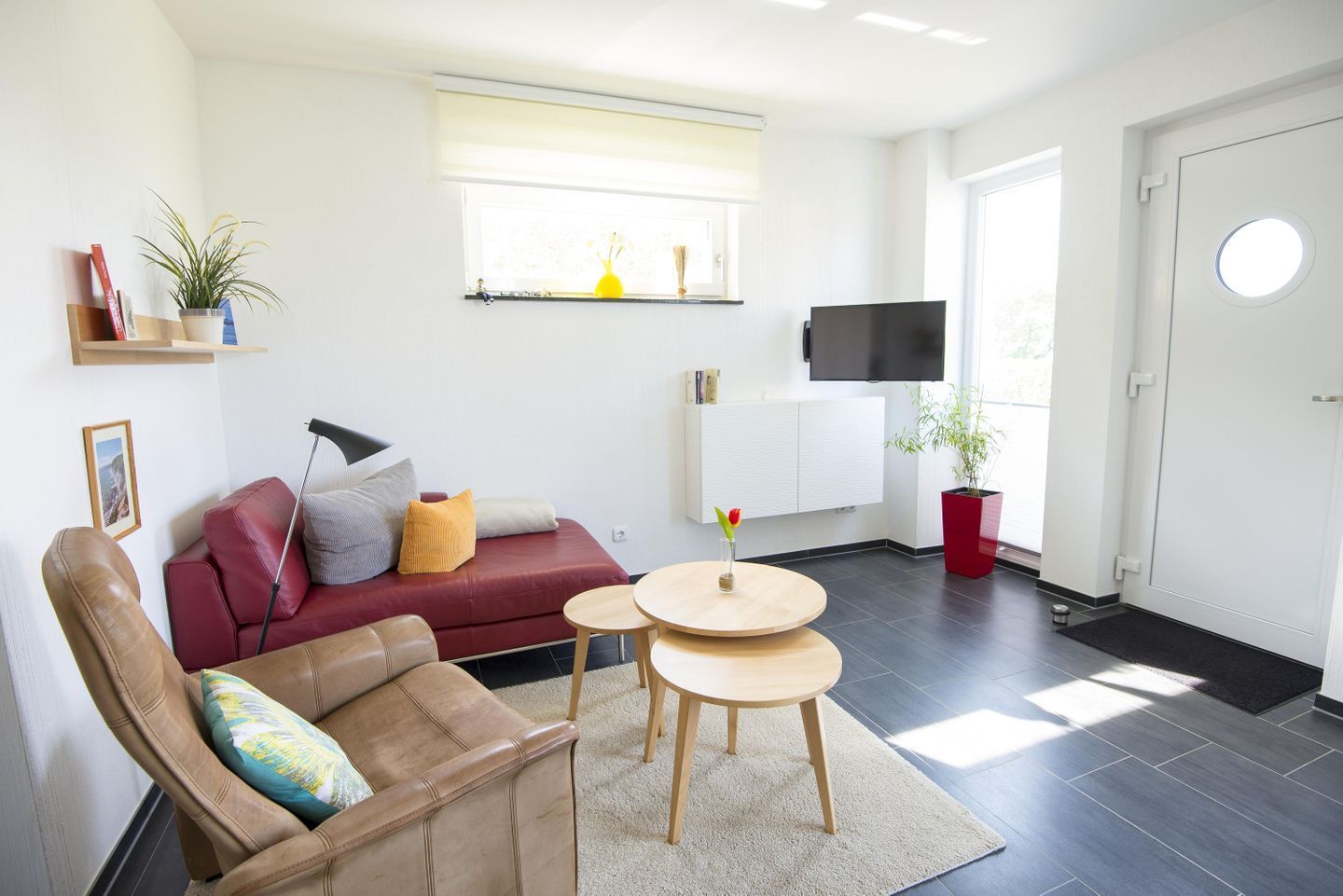 A modern living room with a red sofa, a brown leather chair, and a small wooden table. There is a TV mounted on the wall, a window with blinds, and a potted plant.