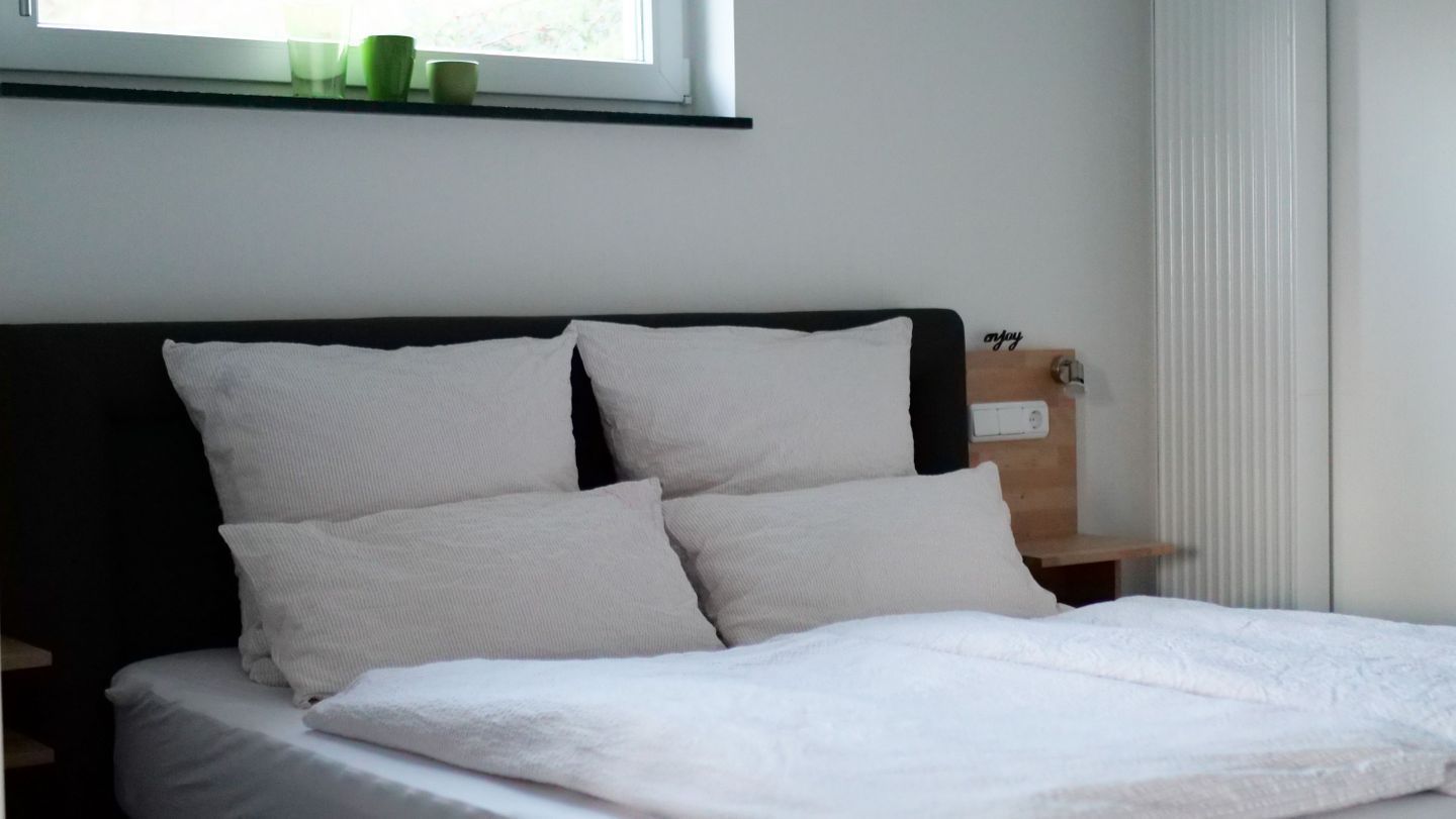 Ein ordentlich gemachtes Bett mit weißen Leinen und Kissen, ein Nachttisch mit einer kleinen Lampe und ein Fenster mit Blick auf Grünflächen.