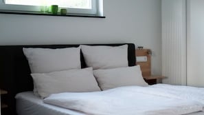 Un lit bien fait, avec du linge de lit blanc et des oreillers, une table de chevet avec une petite lampe, et une fenêtre donnant sur un paysage verdoyant.