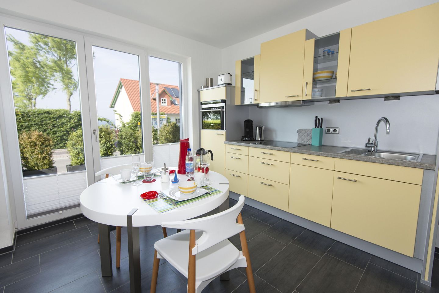 Une cuisine moderne avec des meubles de couleur jaune, une table à manger blanche et une vue sur une maison, visible à travers de grandes fenêtres.