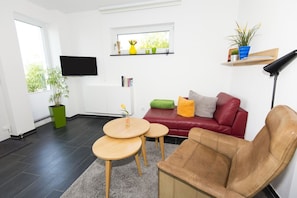 Un salon moderne avec un canapé en cuir rouge, un fauteuil en cuir marron, une table basse en bois et une petite table ronde. Un téléviseur est fixé au mur, il y a une fenêtre donnant sur un espace verdoyant, ainsi qu’une étagère avec des plantes et des livres.