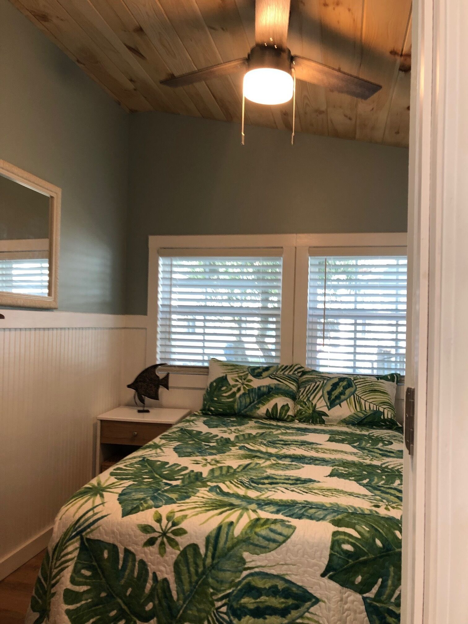 A bedroom with a wooden ceiling, a bed with a tropical leaf-patterned bedspread, a nightstand with a lamp, and a window with blinds.