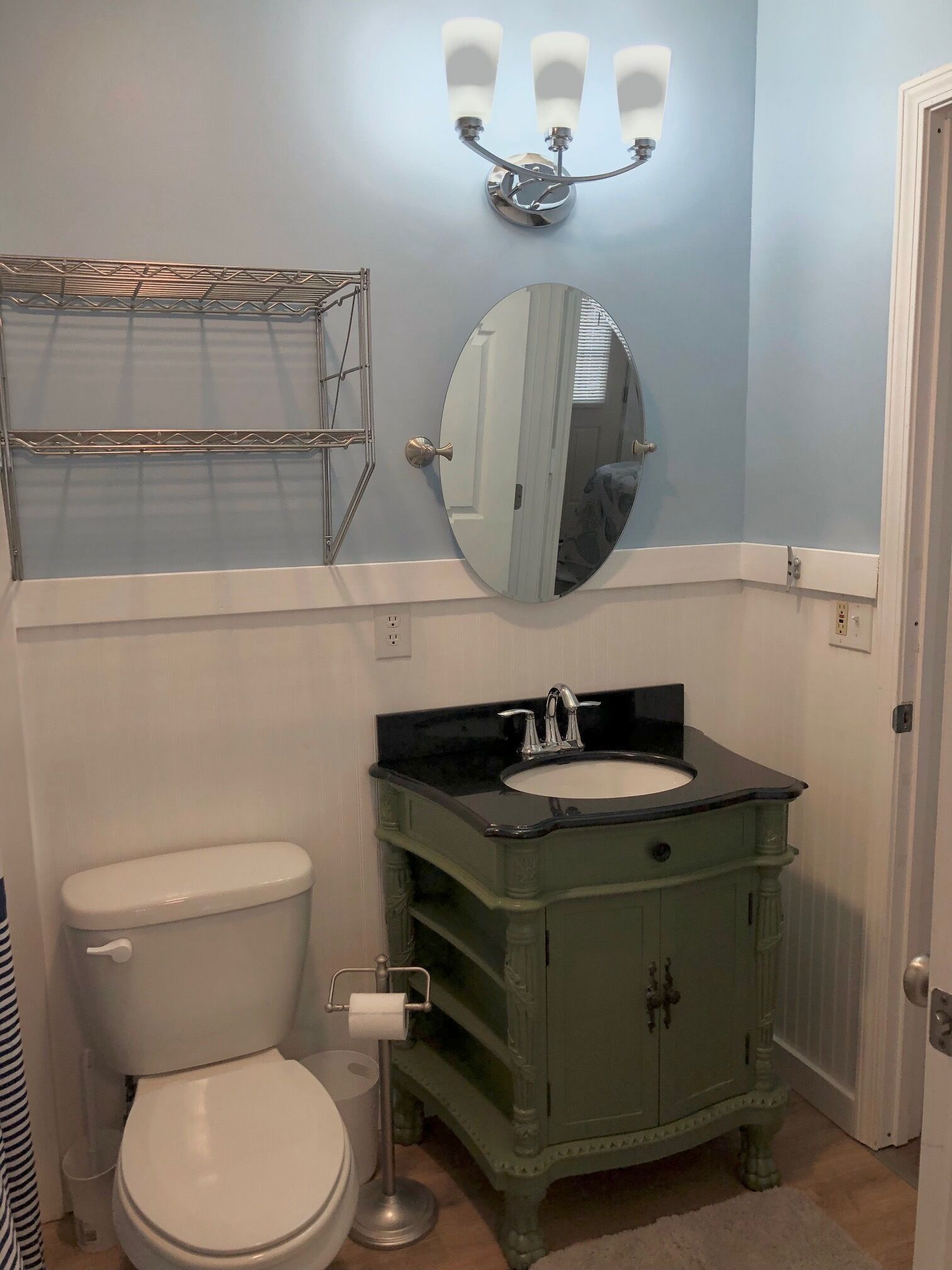 A bathroom with a white toilet, a green vanity with a black countertop, a round mirror, and a wall-mounted light fixture.