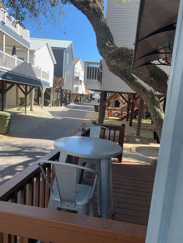 A street view with a wooden deck, a table, and chairs. There are buildings on either side of the street.