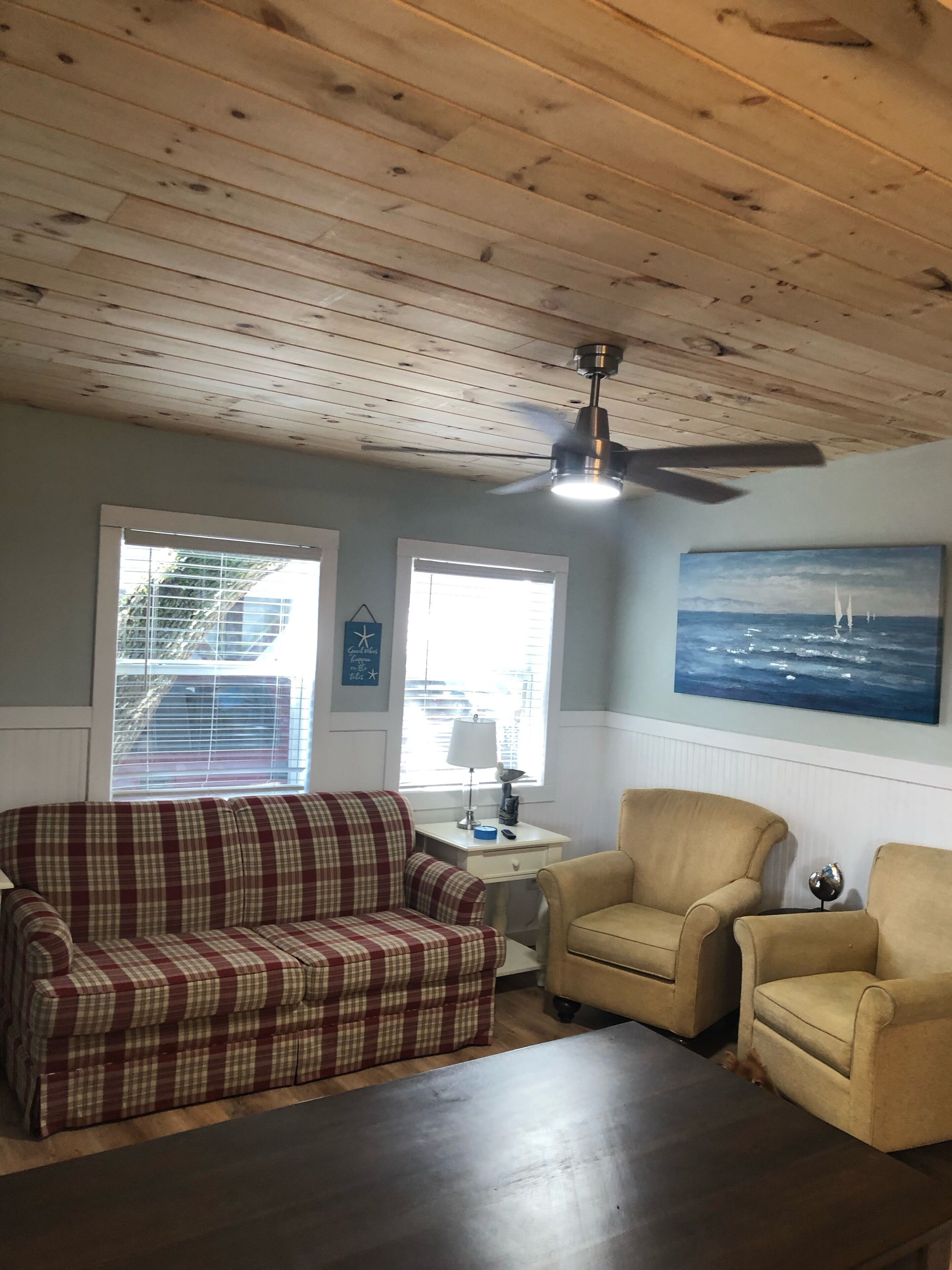 A living room with a plaid sofa, two armchairs, a wooden ceiling, a ceiling fan, a painting of sailboats, and a window with blinds.