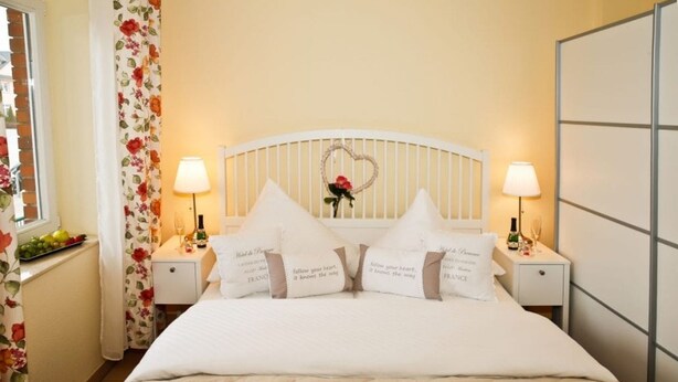 Una cama bien hecha con ropa blanca y almohadas en forma de corazón, un cabecero blanco y dos lámparas de noche.