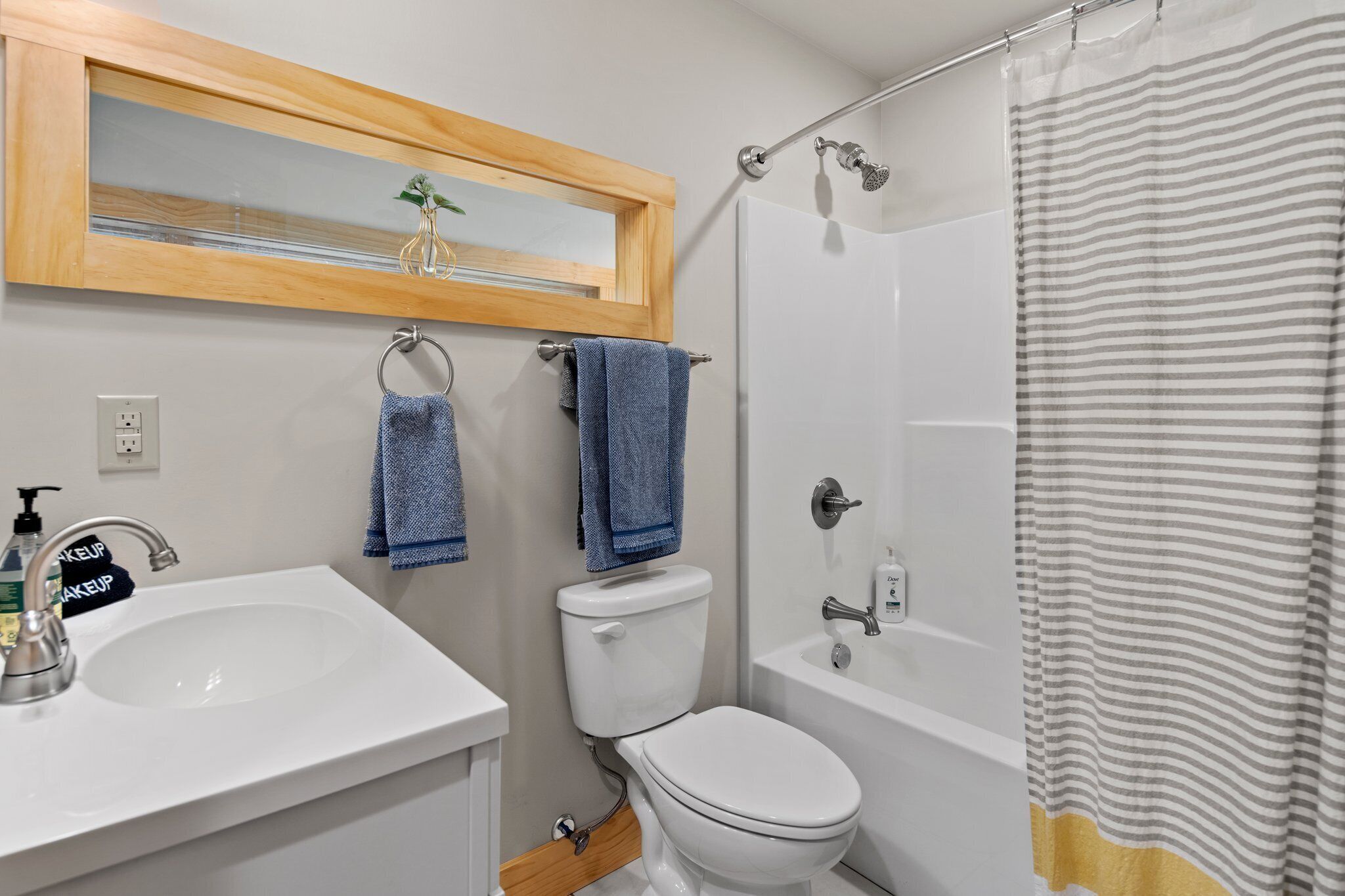 Un baño con lavamanos blanco, inodoro y ducha. Hay un espejo con marco de madera sobre el lavamanos y una cortina de ducha a rayas.