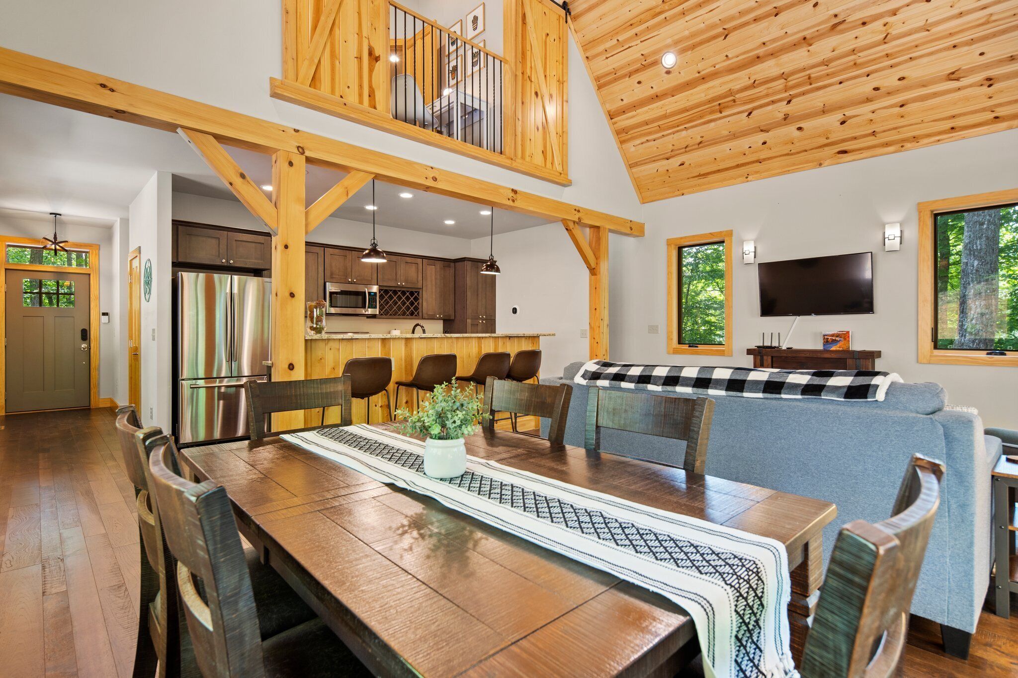 Un vaste espace de salle à manger avec une table et des chaises en bois, une cuisine à l’arrière-plan et une grande fenêtre donnant sur les arbres.
