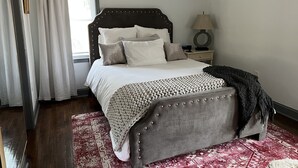 Una cama bien hecha con cabecera, una lámpara de noche y un tapete rojo estampado.